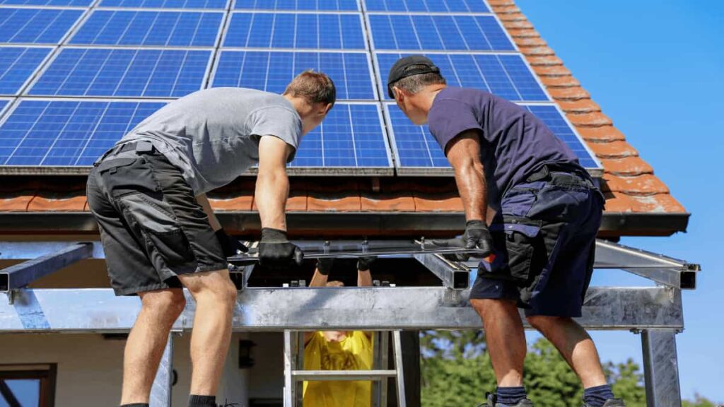 Twee werkmannen installeren het dak van een terrasoverkapping met zonnepanelen op een zonnige dag, terwijl een derde werkman op de achtergrond zichtbaar is.